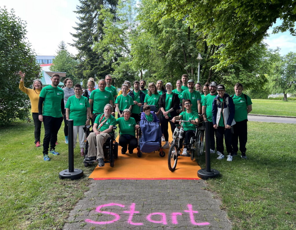 Eine Gruppe von Menschen, einige in Rollstühlen, posiert im Freien auf einem mit rosa Kreide markierten START-Weg. Die meisten tragen passende grüne Hemden, und im Hintergrund sind Bäume und Gras zu sehen.