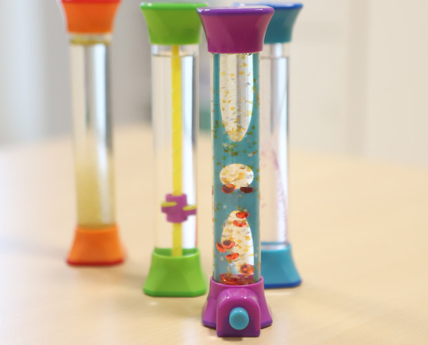 Vier farbenfrohe Timer mit Flüssigkeiten in verschiedenen Formen und Farben stehen auf einer hellen Oberfläche. Die Timer enthalten schwimmende Blasen und Öl, was einen beruhigenden visuellen Effekt erzeugt.