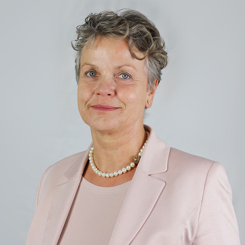 Eine Frau mit kurzen, lockigen grauen Haaren, die einen hellrosa Blazer, ein passendes Oberteil und eine Perlenkette trägt, posiert vor einem schlichten hellgrauen Hintergrund und lächelt sanft.