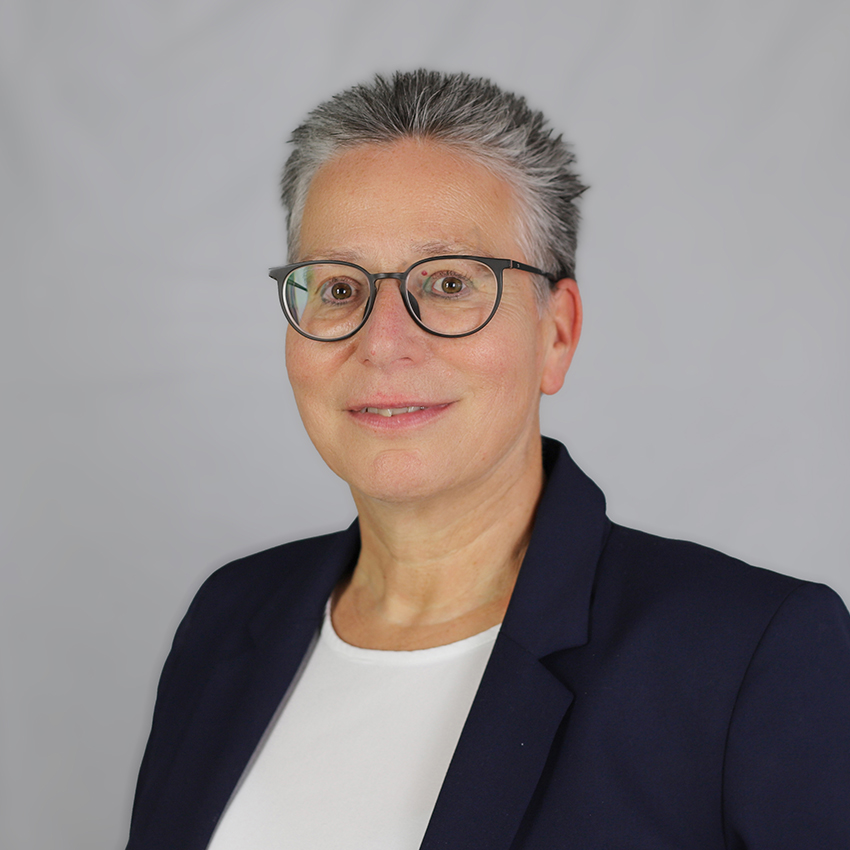 Eine Person mit kurzen grauen Haaren, Brille und einem marineblauen Blazer über einem weißen Hemd lächelt in die Kamera vor einem schlichten hellgrauen Hintergrund.