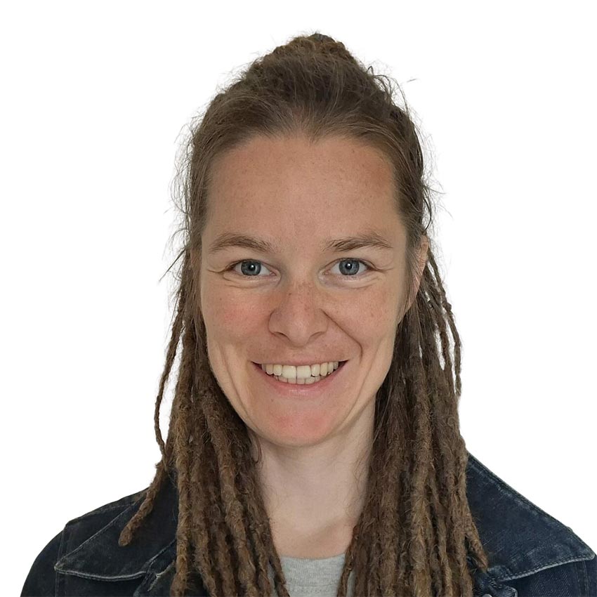 Lächelnde Frau mit langen braunen Dreadlocks, die eine dunkle Jeansjacke über einem hellgrauen Hemd trägt, fotografiert vor einem schlichten weißen Hintergrund.