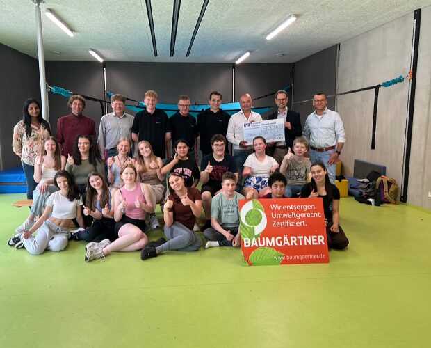 Eine Gruppe von Personen Erwachsenen und Kindern in einer Turnhalle bei einem Gruppenbild ein Check wird hochgehalten ein Schild der Spende