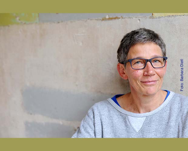 Frau im Portgrait Fotografier vor einer Wand mit Grobputz Sie trägt eine Blaue Hornbrille, ein graues Oberteill die Haare sind evbenfalls in grauer Kurzharfrisur