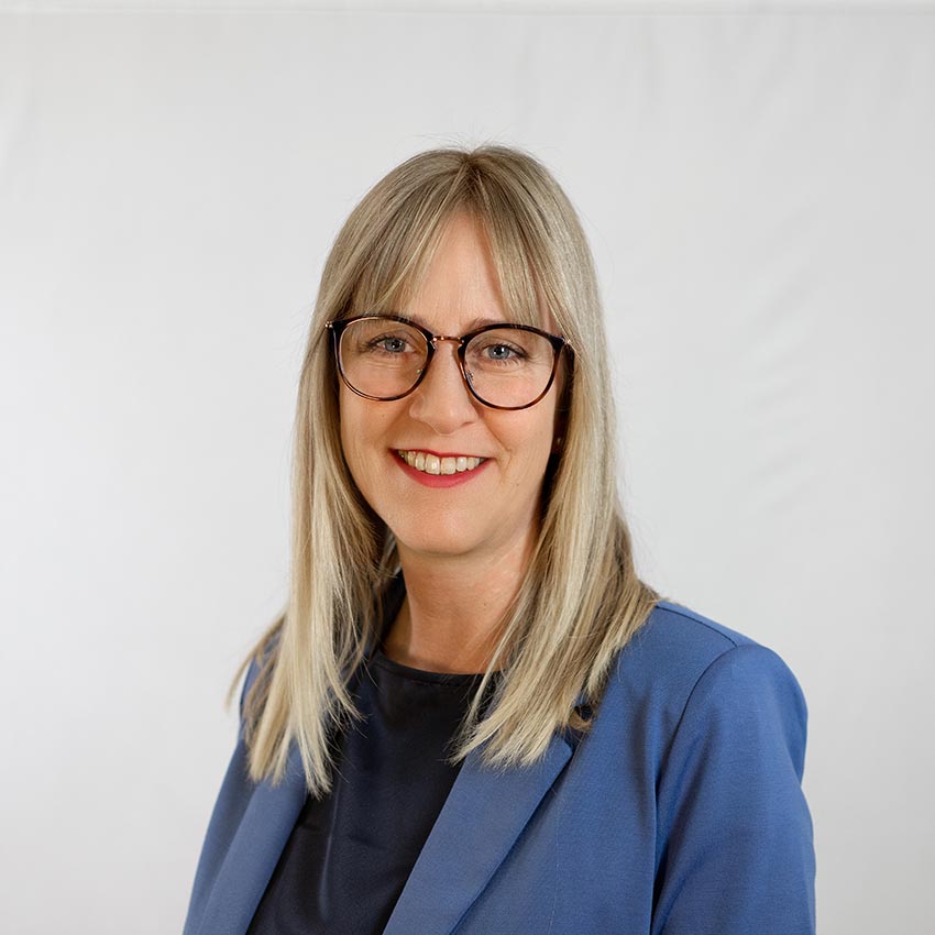 Eine Frau mit Brille und glattem, langem, blonden Haar und einem blauen Oberteil lächelt in die Kamera. Der Hintergrund ist einfarbig und hell.