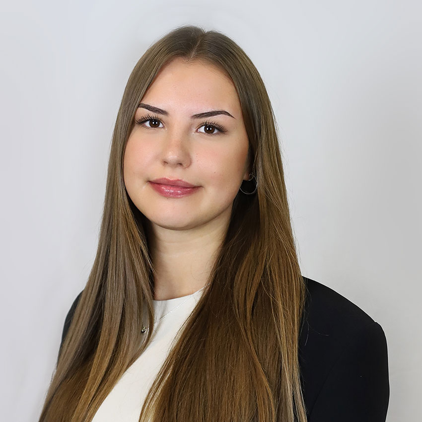 Eine junge Frau mit braunen langen Haaren und braunen Augen, trägt Ohrringe, eine blaue Blaser-Jackeund ein weißes Oberteil. Sie lächelt. Es handelt sichum ein Portrait