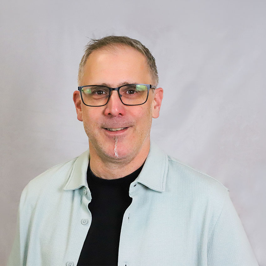 Ein Mann mit Brille lächelt in die Kamera Er hat einen schmalen Kinstrichbart und trägt eine mindgrüne West mit schwarzem T-shirt