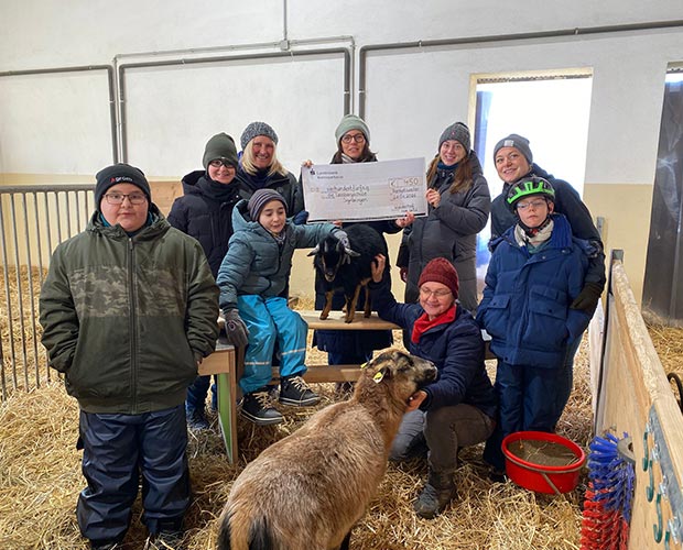 Auf dem Bauernhof: Eine Personengruppe mit Kindern in einem Stall halten einen Scheck und stehen auf Stroh im ist Fodderfgrund ist eine Ziege zu sehen