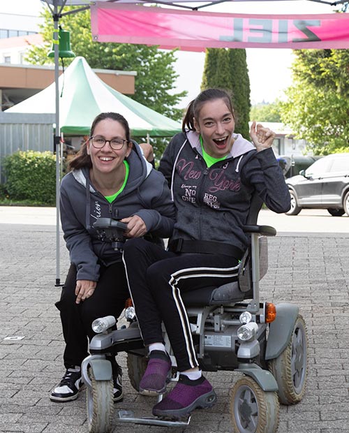 Zwei junge Frauen, eine davon im Rollstul sitzend und die andere neben Ihr kniend lachen in die Kamera unter einem Pavilion vor einem Ziel Banner