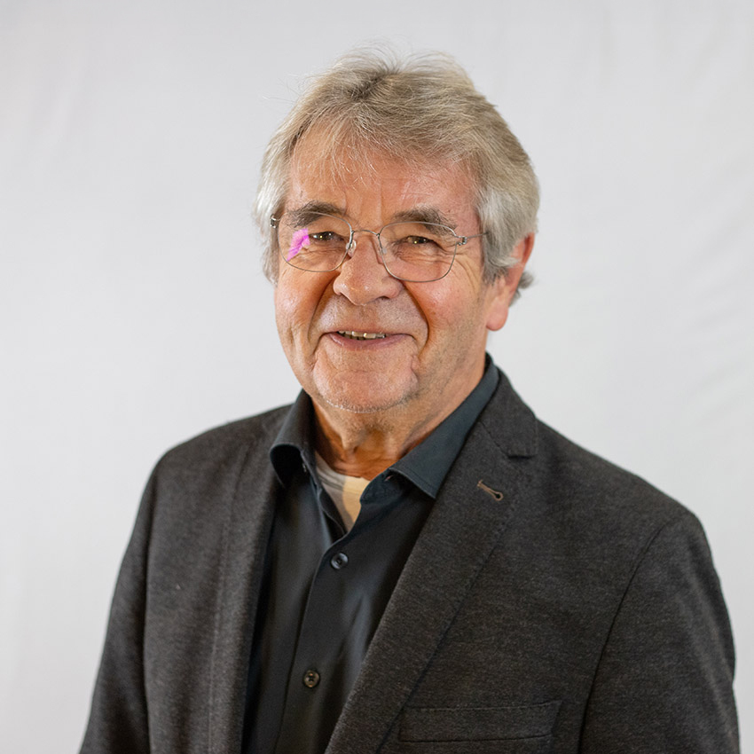 Ein älterer Mann mit grauem Haar und Brille, der einen dunklen Blazer über einem schwarzen Hemd trägt, lächelt in die Kamera vor einem schlichten hellen Hintergrund.